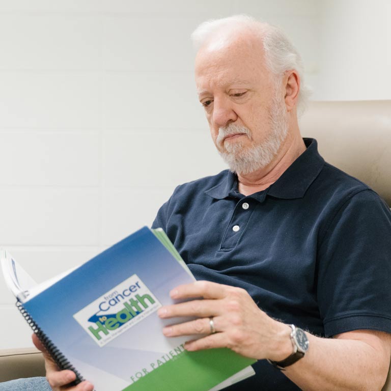 older man sitting in chair reading a book that reads from cancer to health